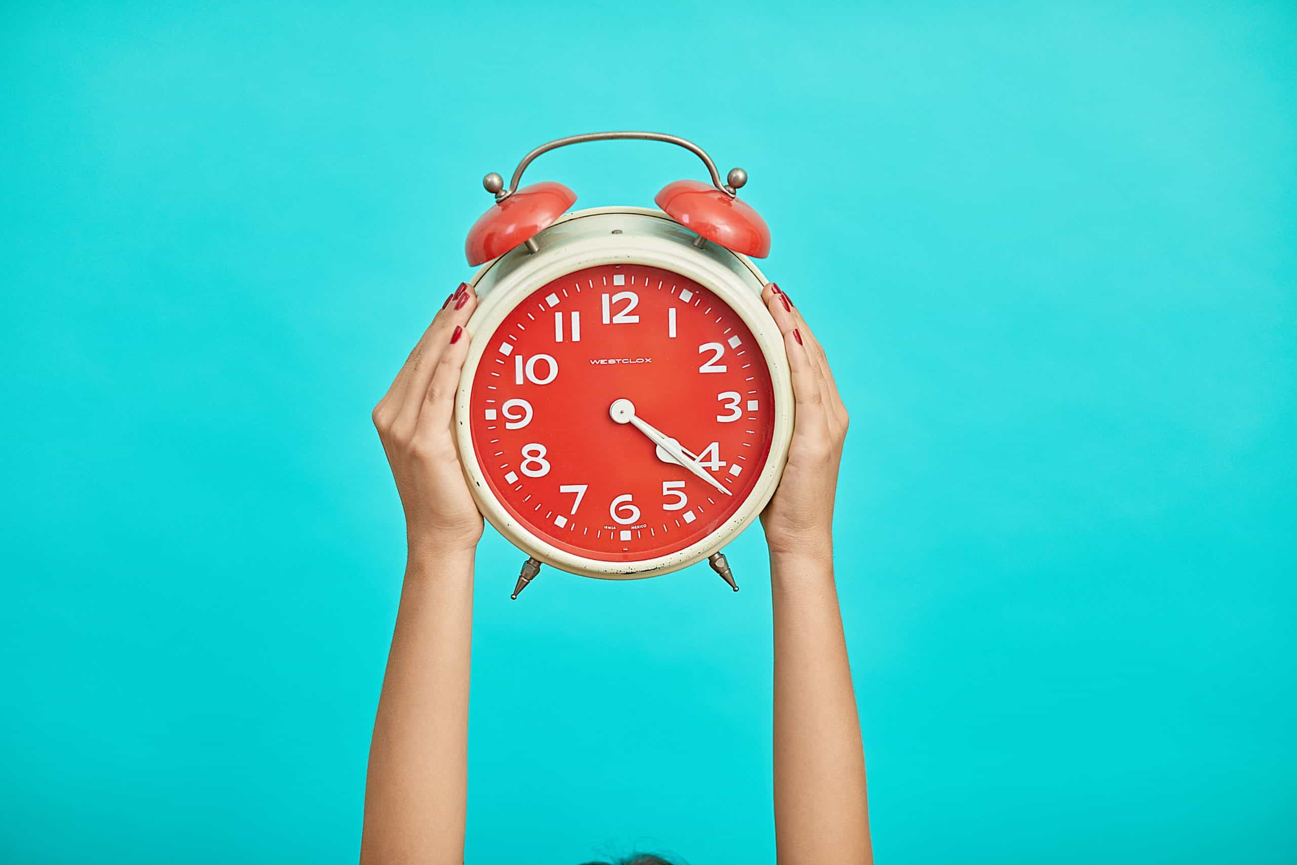 Bright blue background with two hands holding up a red alarm clock.