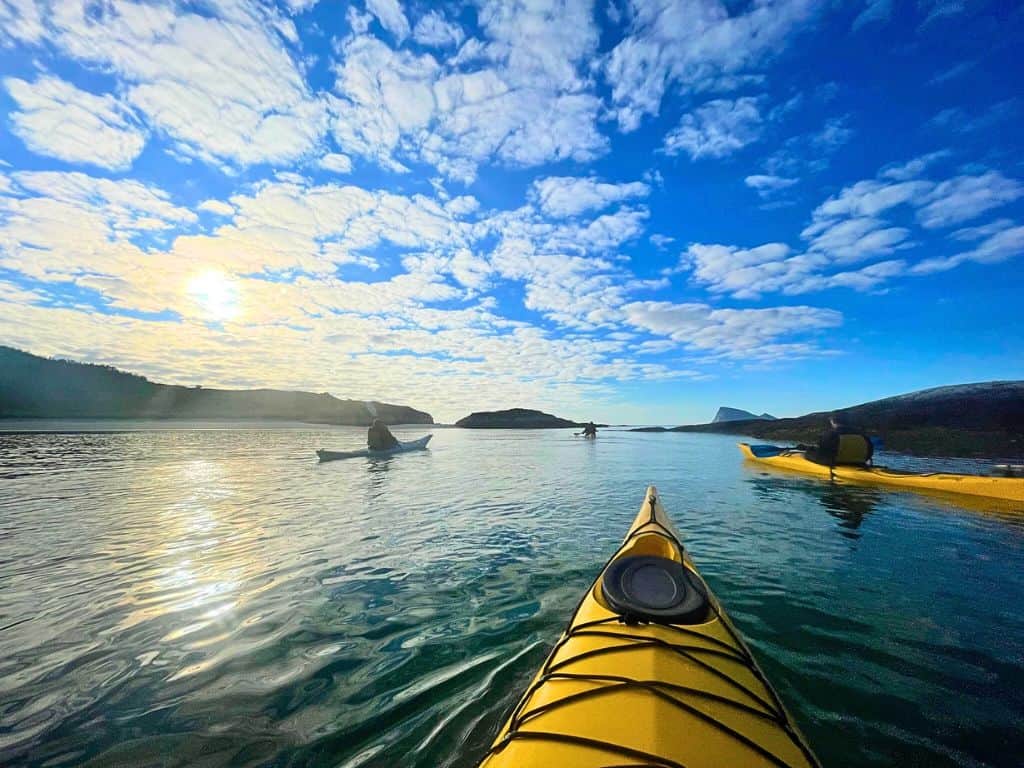 Yellow kayaks on the water with a blue sky and some clouds.  There are four kayaks and some mountains nearby.  The sun is behind the cloud as it is starting to look like it's setting.