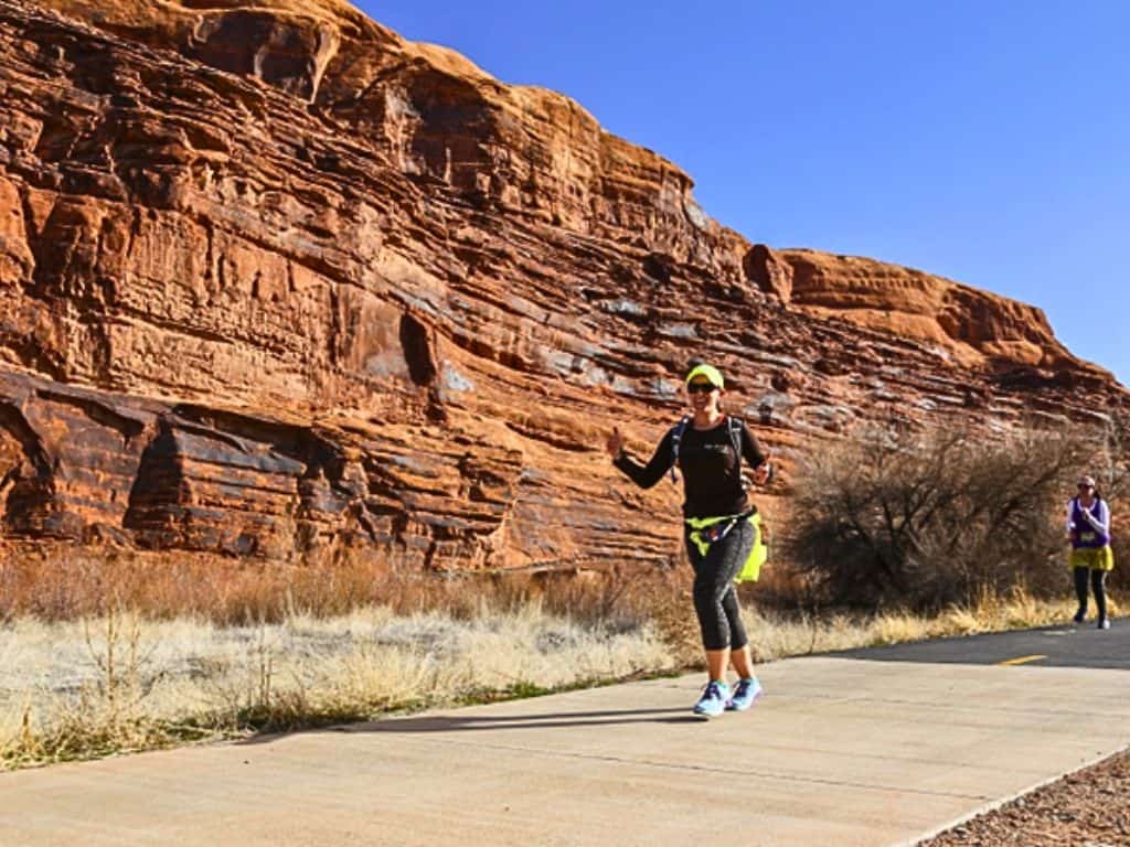 Woman running on the paved sidewalk.  Red rocks frame the background with a blue sky.