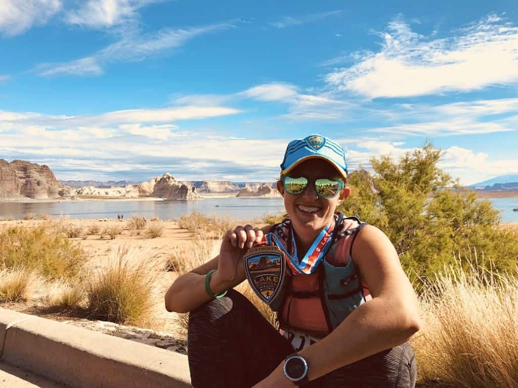 Lucy, the author, sitting on the curb holding her Lake Powell finisher medal up.  She's smiling to the camera.  In the background there's desert brush and a beautiful lake with canyon mountains in the distance.