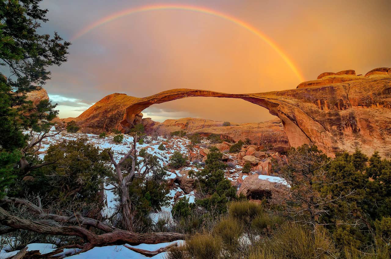 Stunning view of Arches National Park.  There's a little snow and greenery in the foreground.  In the background there's the famous long thin arch connecting two larger rocks.  There's a rainbow in the soft velvety sky.
