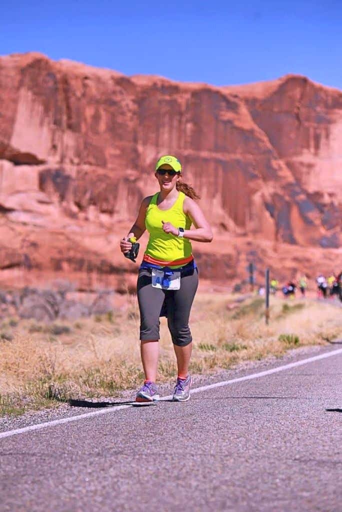 Woman running wearing a racing bib, sunglasses, and hat.  There are other runners blurred running in the distance.  The race is on the road and there's red rock canyons behind.