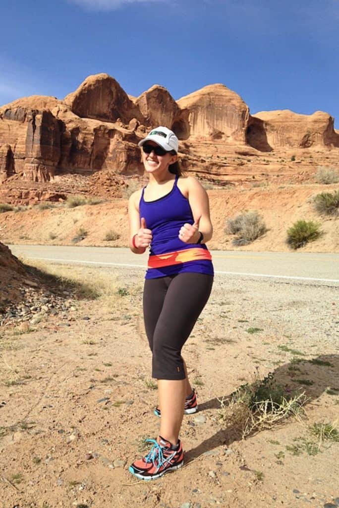 The author, Lucy, posing in her running gear giving a double thumbs up.  In the background is a road with desert scenery and orange canyons.
