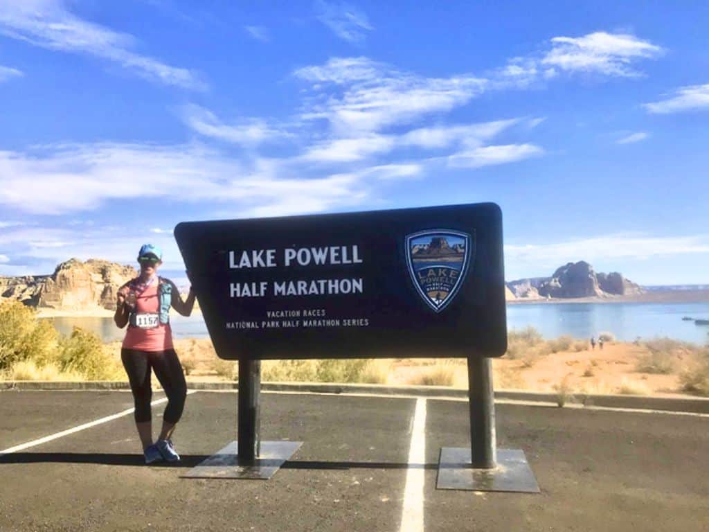 The author, Lucy, posing with a Lake Powell half marathon sign.  She's wearing her racing bib and holding her medal up.  In the background there's a lake, distant canyons, and a blue sky