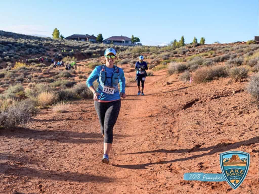 Official race photo with the Lake Powell logo in the bottom right corner.  Lucy, the author, is running on an orange red trail heading toward the camera.  She's smiling, wearing her hat, sunglasses, and racing bib.