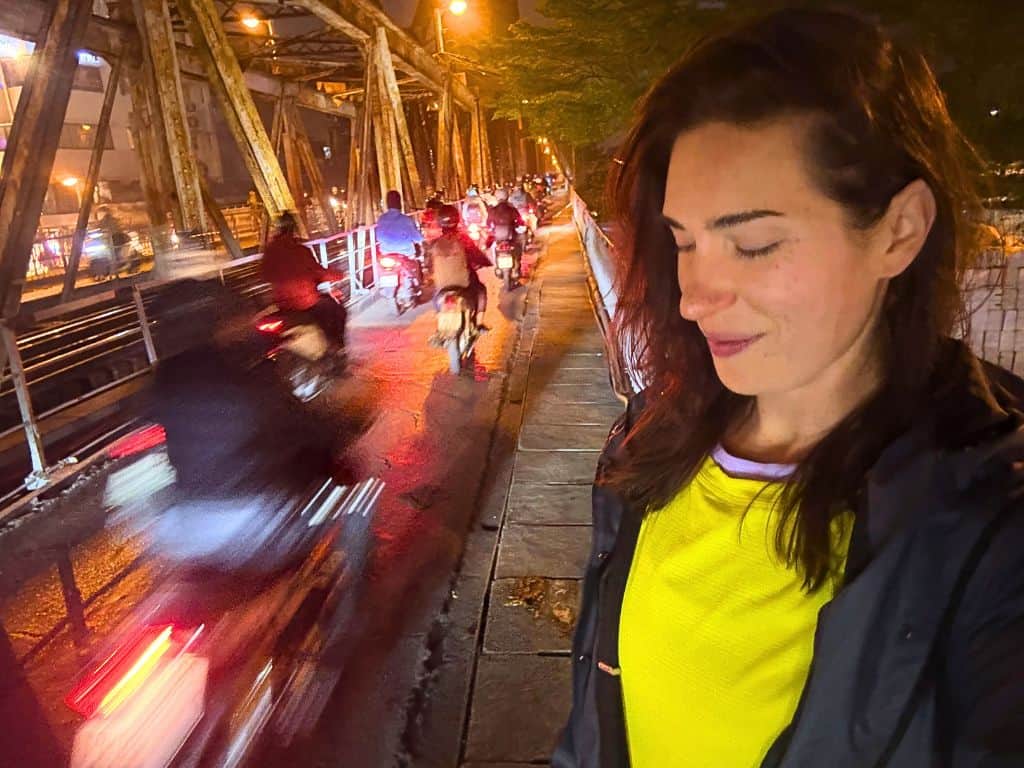 Heidi standing next to the blurred, busy street of the scooters rushing by on the bridge