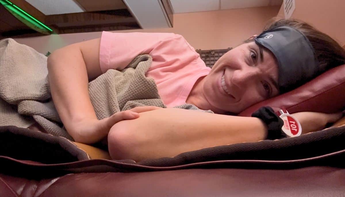 Heidi getting ready to sleep overnight at the King Spa Chicago. She's wearing a face mask above her eyes, has a blanket, and is wearing her uniform laying on a bed. She also has the King Spa wristband with her barcode and number on it.