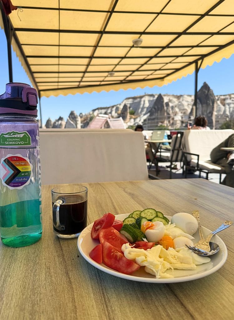 Plate of food with tomatoes, cucumber, hard boiled eggs, and cheese. There's a coffee mug and water bottle on the table. View is from a hostel terrace in Cappadocia.
